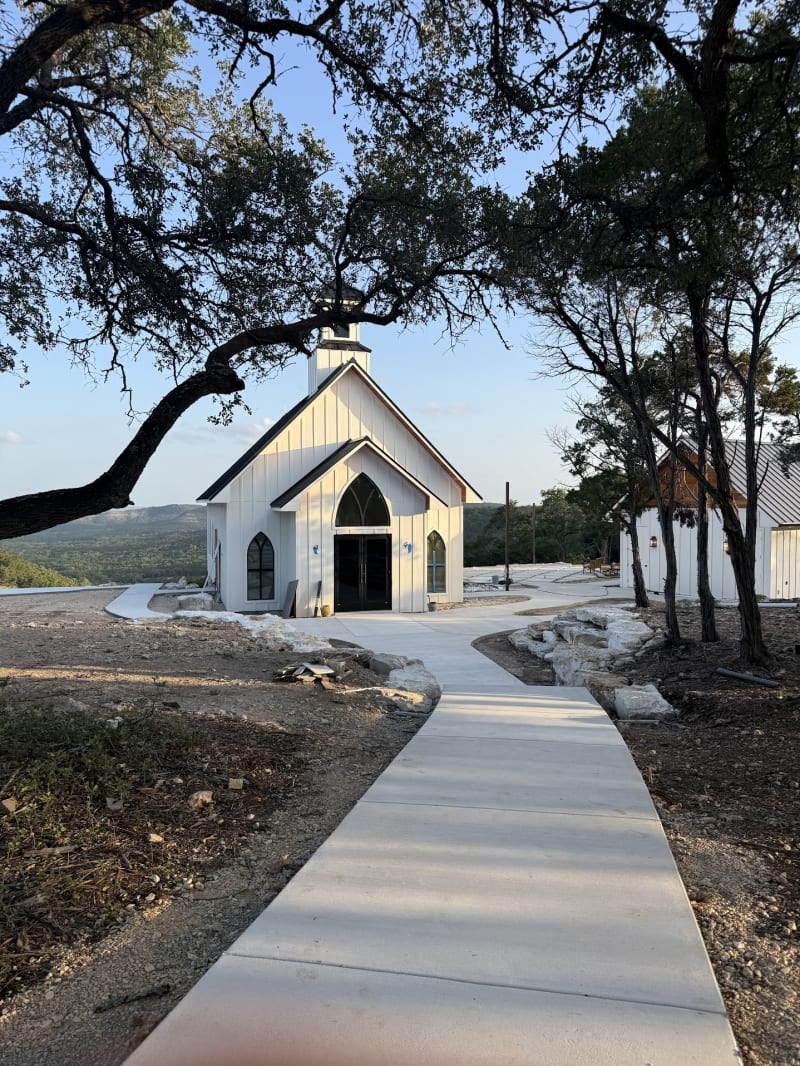 Concrete path leading beneath oak trees to The Chapel at Wimberley, a white wedding chapel on a Texas Hill Country hilltop at golden hour